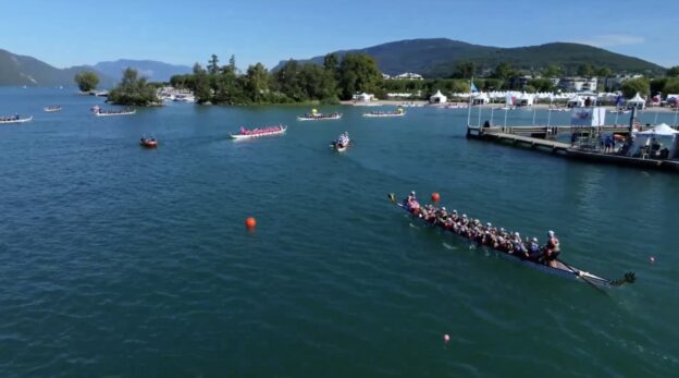 A photo showing a lake with mountains in the background. In the foreground are several "dragon boats", which are large, canoe-like boats with 20 people in them paddling. Behind the boats there is a beach with tents and people visible on it.
